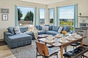 A modern living room with a sectional sofa, dining table, chairs, and large windows offering a view of greenery and buildings.
