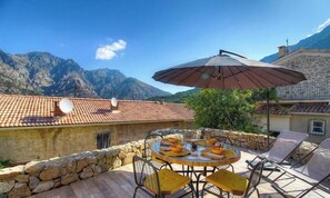 Eine Terrasse mit einem runden Tisch und Stühlen, einem großen Sonnenschirm und einer Bergkulisse im Hintergrund.