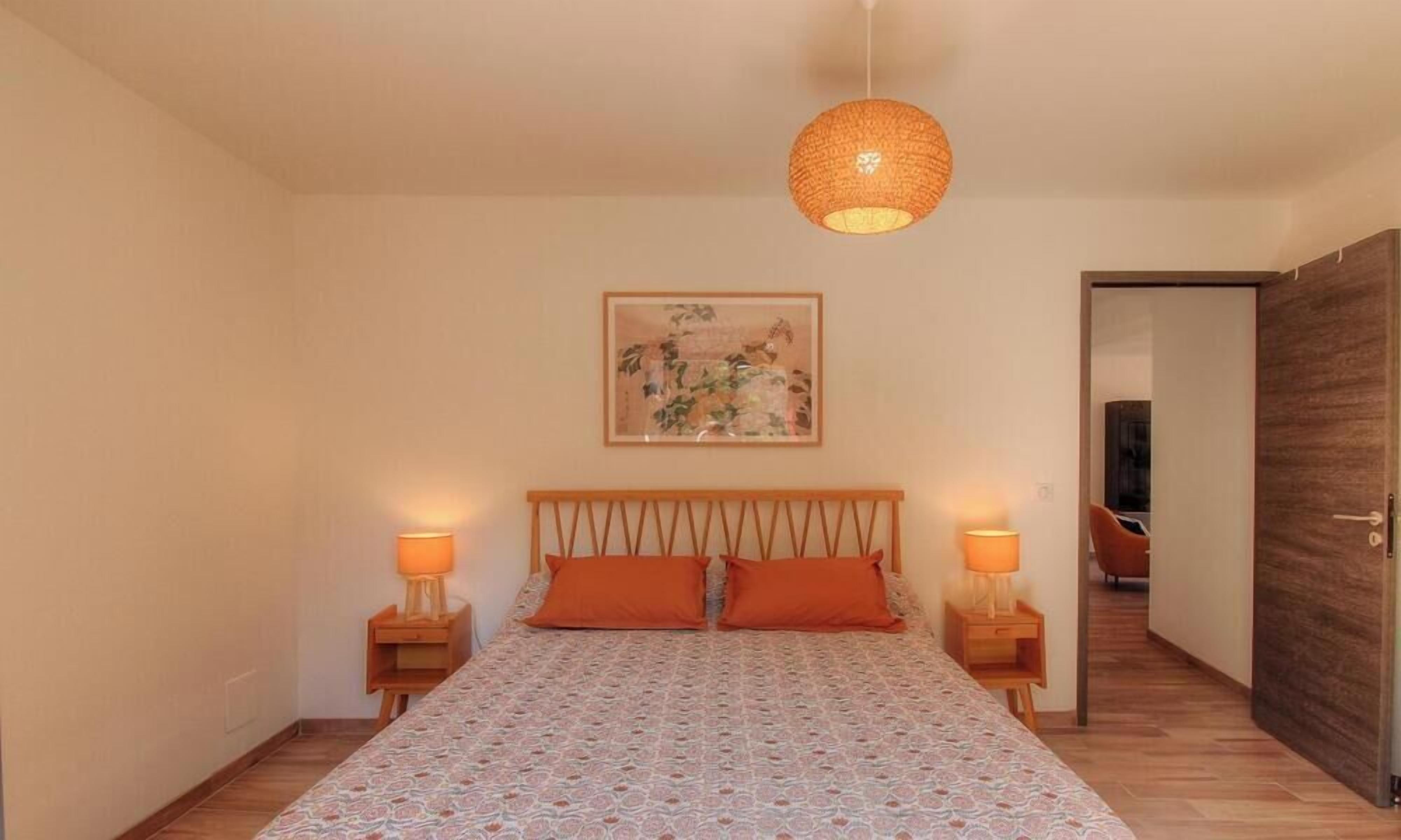 Une chambre à coucher avec un lit, deux lampes de chevet, un luminaire suspendu, un tableau encadré au mur et une porte donnant sur une autre pièce.