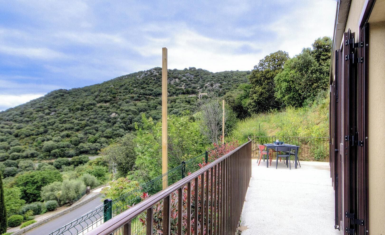 A balcony with a view of a hillside, a table with chairs, and a building with a door.