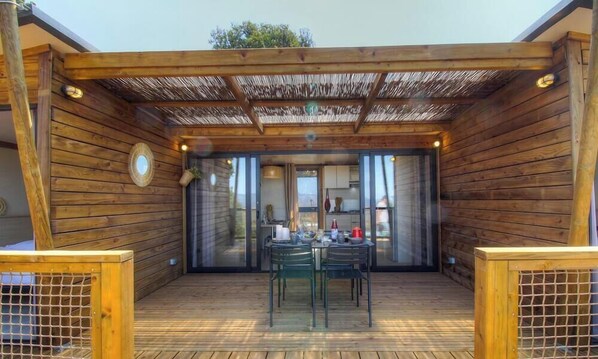 Una terraza de madera con mesa y sillas, una pérgola y vistas al interior.