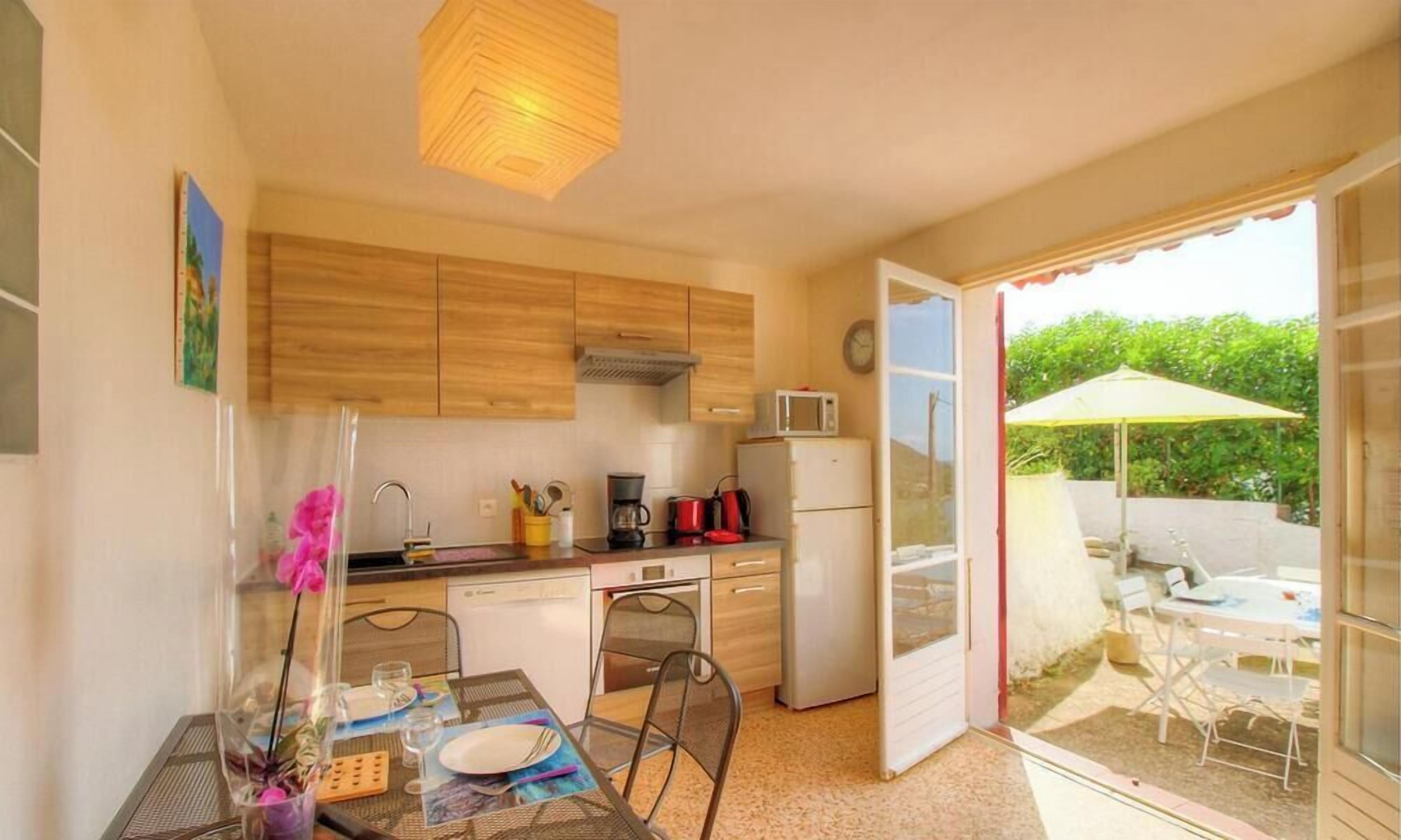 Cocina compacta con armarios de madera, microondas y refrigerador. Cuenta con comedor con mesa y sillas, y un patio con sombrilla.