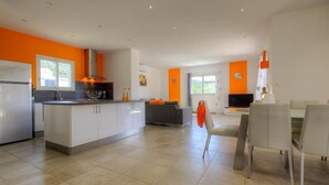 Una cocina moderna con una isla central, una zona de comedor con mesa y sillas, y una sala de estar con sofá y televisor.