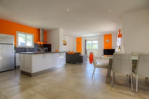 Eine moderne Küche mit Kochinsel, ein Essbereich mit Tisch und Stühlen sowie ein Wohnzimmer mit Sofa und Fernseher.