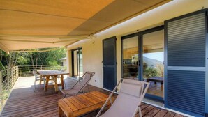 Une terrasse en bois avec une table et des chaises, une porte-fenêtre coulissante et une vue sur la verdure.