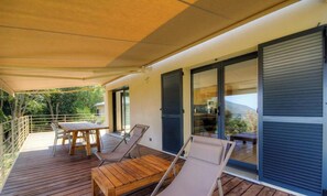 A wooden deck with a table and chairs, a sliding glass door, and a view of greenery. - Santa Maria - SISCO - Gîte (SISCO)
