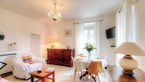 A living room with a white sofa, a wooden cabinet, a dining table with chairs, a chandelier, and a TV mounted on the wall. - Le 22401 - PETRETO BICCHISANO (PETRETO BICCHISANO)