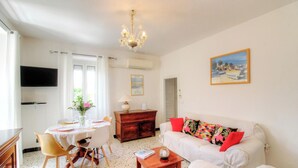 A living room with a white sofa, a dining table with chairs, a wooden cabinet, a chandelier, and a painting on the wall. - Le 22401 - PETRETO BICCHISANO (PETRETO BICCHISANO)