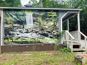 Una estructura de madera con un mural de cascada, un cartel que dice '¡Bienvenidos Campistas!' y un letrero de 'Propiedad Privada. Prohibido el Paso'.
