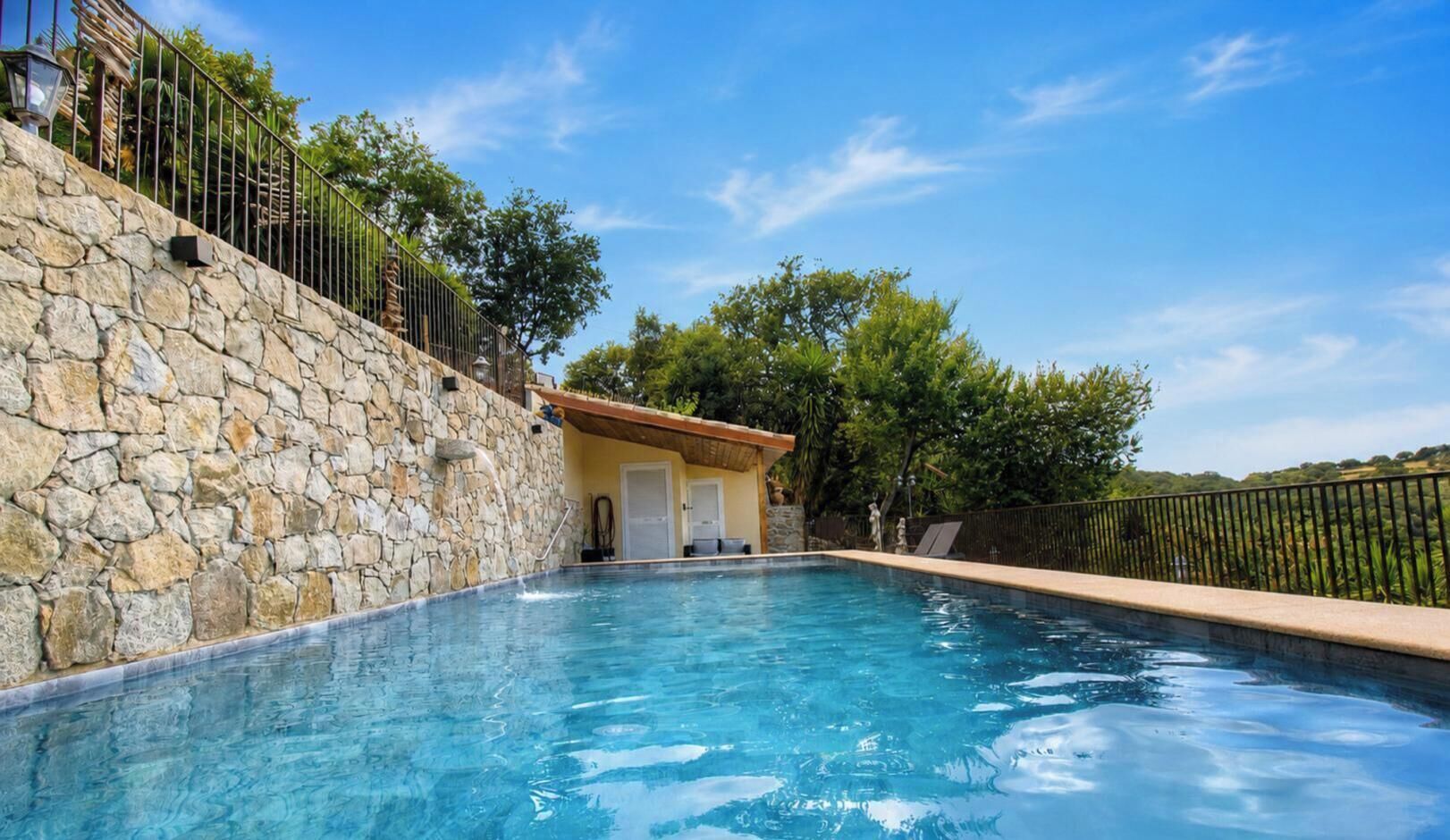 A swimming pool with stone walls, a small building with a tiled roof, and a metal fence.