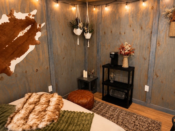 A room with a wooden wall, a bed with a fur throw, a small black table with a vase, and hanging macramé planters.