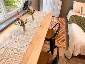 A wooden dining table with a macrame runner, two chairs, a bed with a green blanket, and a window with a view of greenery.