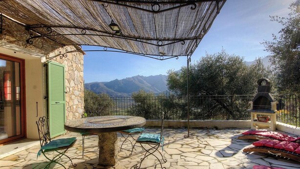 Eine Terrasse im Freien mit Steinboden, einem runden Tisch, einem Metallschirm und einem Grill.