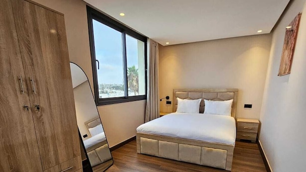 A modern bedroom with a large bed, wooden wardrobe, and a view through a window. - OCEAN ROOFTOP HOTEL (El Mansouria)