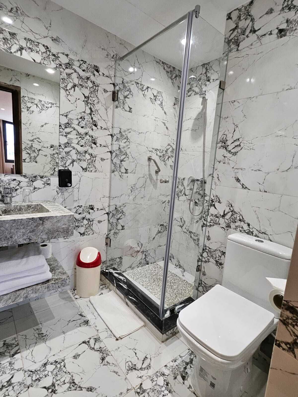 Un baño con ducha de vidrio, paredes de mármol, inodoro blanco y lavamanos con encimera de mármol.