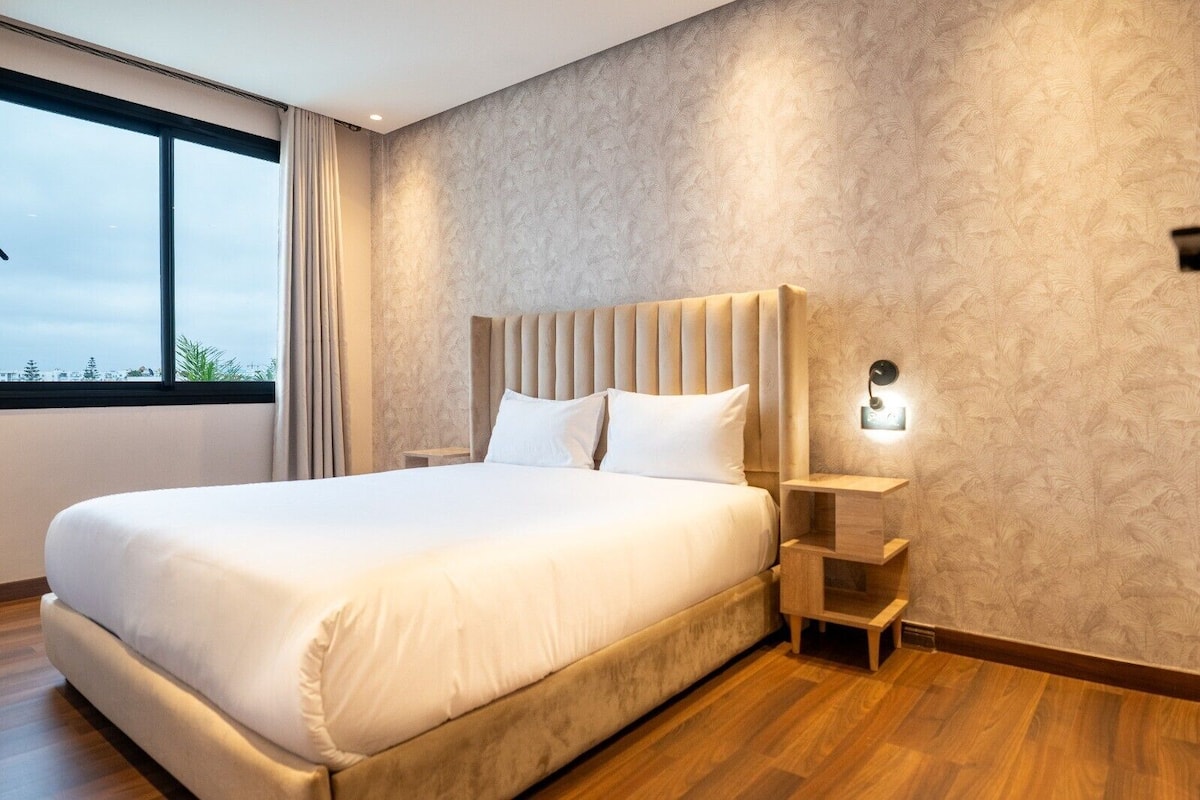 A hotel room with a large bed, a nightstand, a wall-mounted lamp, and a window with curtains.