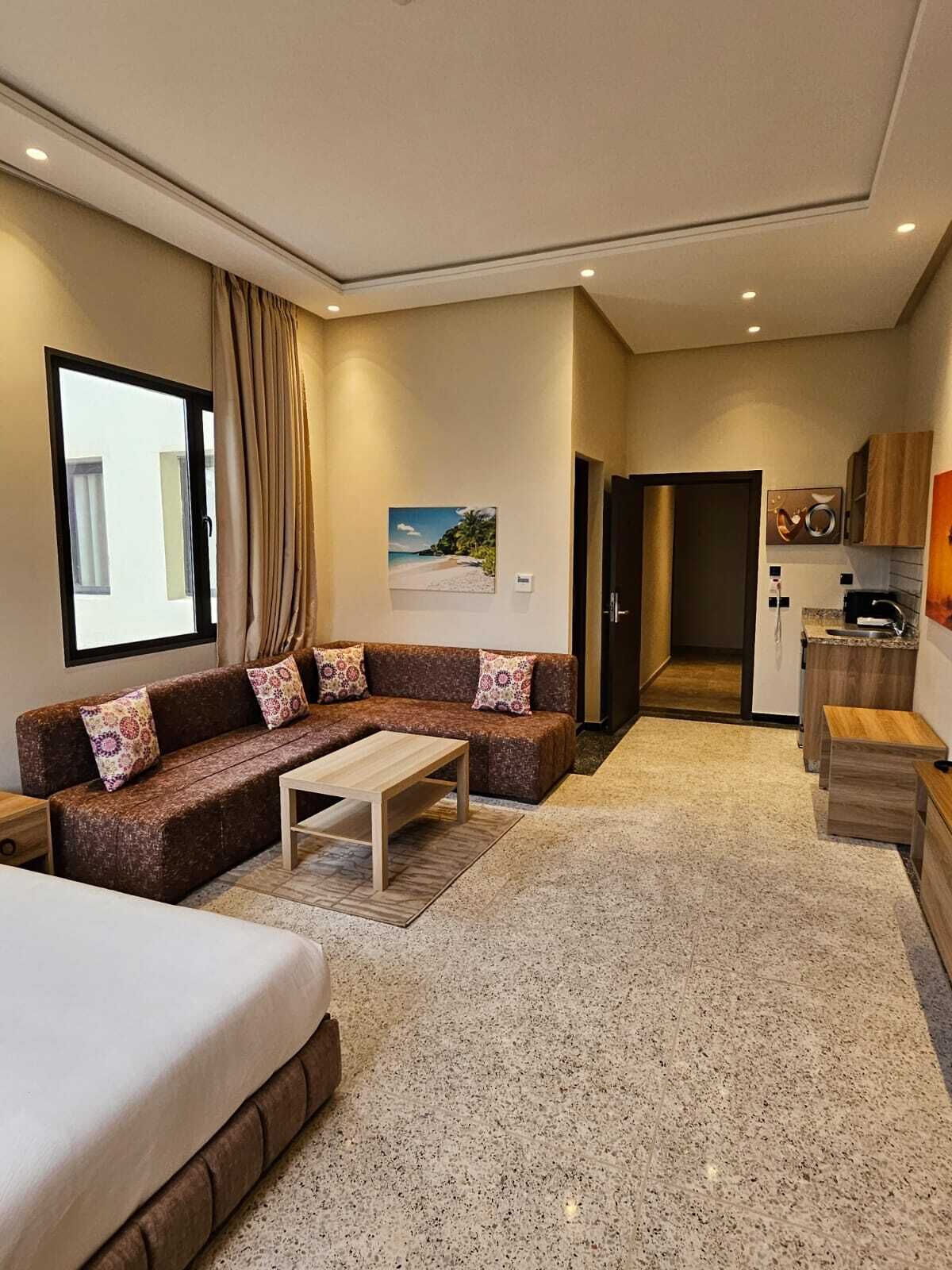 A modern hotel room with a sofa, a small table, a bed, a kitchenette, and a view of the outdoors.
