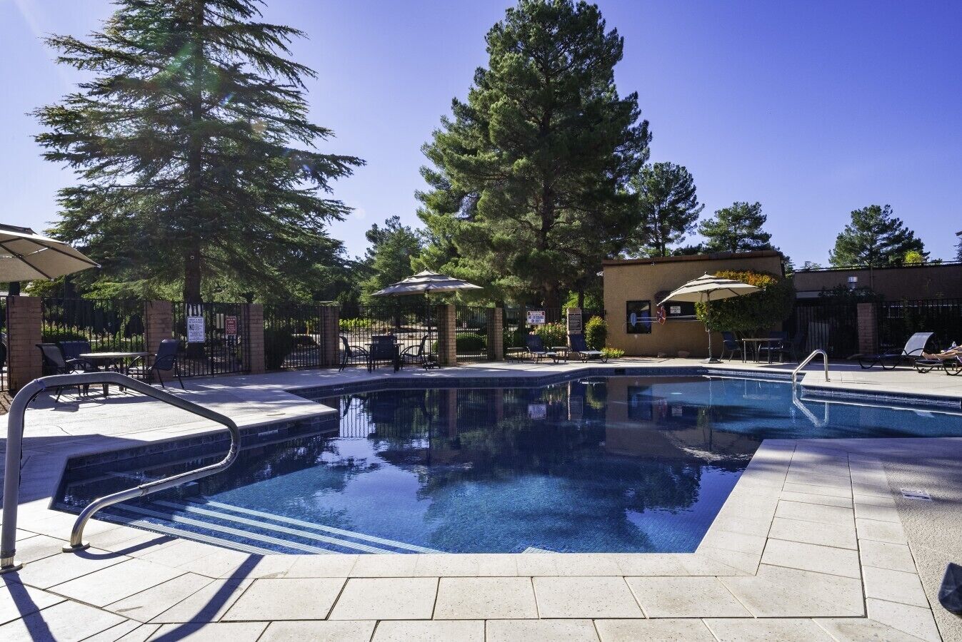 Ein Pool mit Metallleiter, umgeben von Liegestühlen und Sonnenschirmen, mit Bäumen und einem Gebäude im Hintergrund.