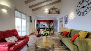 Un salón amplio con un sofá de cuero rojo, un sofá verde y una mesa de centro redonda. Se observa una cocina al fondo con armarios rojos y un comedor.