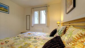Un dormitorio con cama, ventana con cortinas, lámpara de noche y un cuadro en la pared.