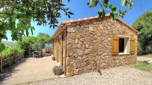 Edificio de piedra con una ventana pequeña y una puerta, rodeado por un camino de grava y follaje.