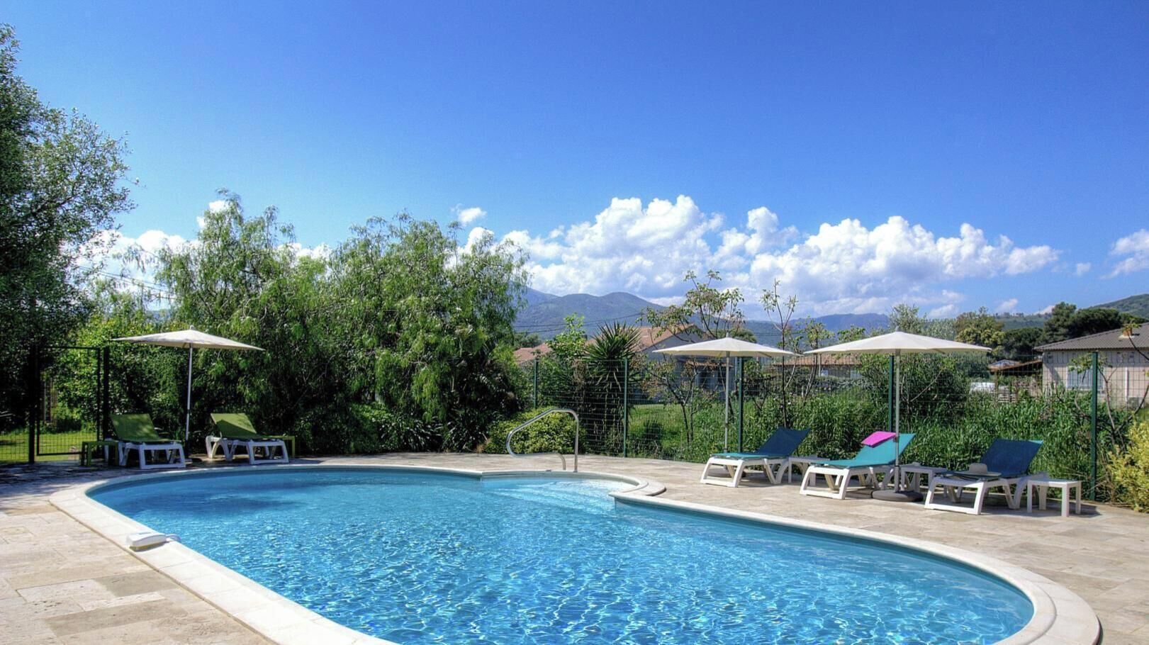 Ein Pool mit Liegestühlen und Sonnenschirmen, umgeben von Grünpflanzen und Bergen im Hintergrund.