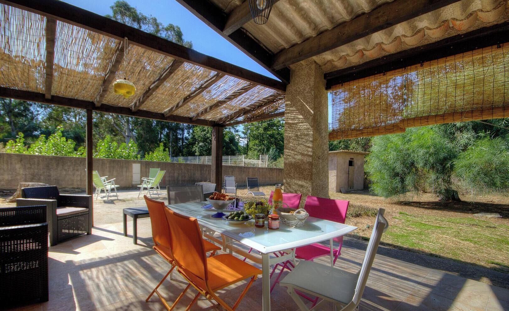 An outdoor patio with a table set for dining, surrounded by chairs, and a covered area with a thatched roof.