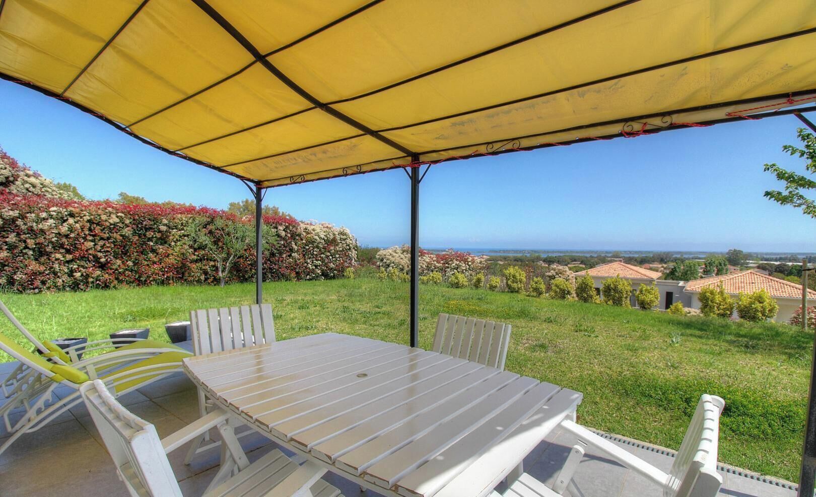 Eine Terrasse im Freien mit Tisch und Stühlen, einem gelben Sonnensegel und Blick auf eine Landschaft mit Gebäuden und blauem Himmel.