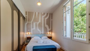 A bedroom with a bed, bedside lamps, a bench, a patterned wall, and a window with a view of greenery. - L'Écrin Lumineux avec double balcon - Welkeys (Arcachon)