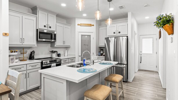 A modern kitchen with white cabinets, stainless steel appliances, a central island with stools, pendant lighting, and a hanging basket with flowers. - The Cozy Ridge Apartment (Edinburg)