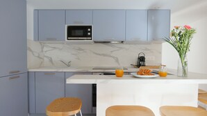 Eine moderne Küche mit grauen Schränken, einer weißen Arbeitsplatte und einer Marmor-Spritzschutzwand. Zwei Holzstühle und ein Tisch mit Frühstücksgebäck sind vorhanden.