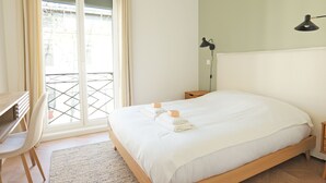 Ein Schlafzimmer mit einem Bett, einem Schreibtisch, einem Stuhl, einem Nachttisch, einer Lampe und Blick durch ein Fenster.