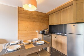 A modern kitchen with wooden cabinetry, a stainless steel refrigerator, and a dining table set with plates and glasses. - Le Cocon du Gollet - Welkeys (Megève)