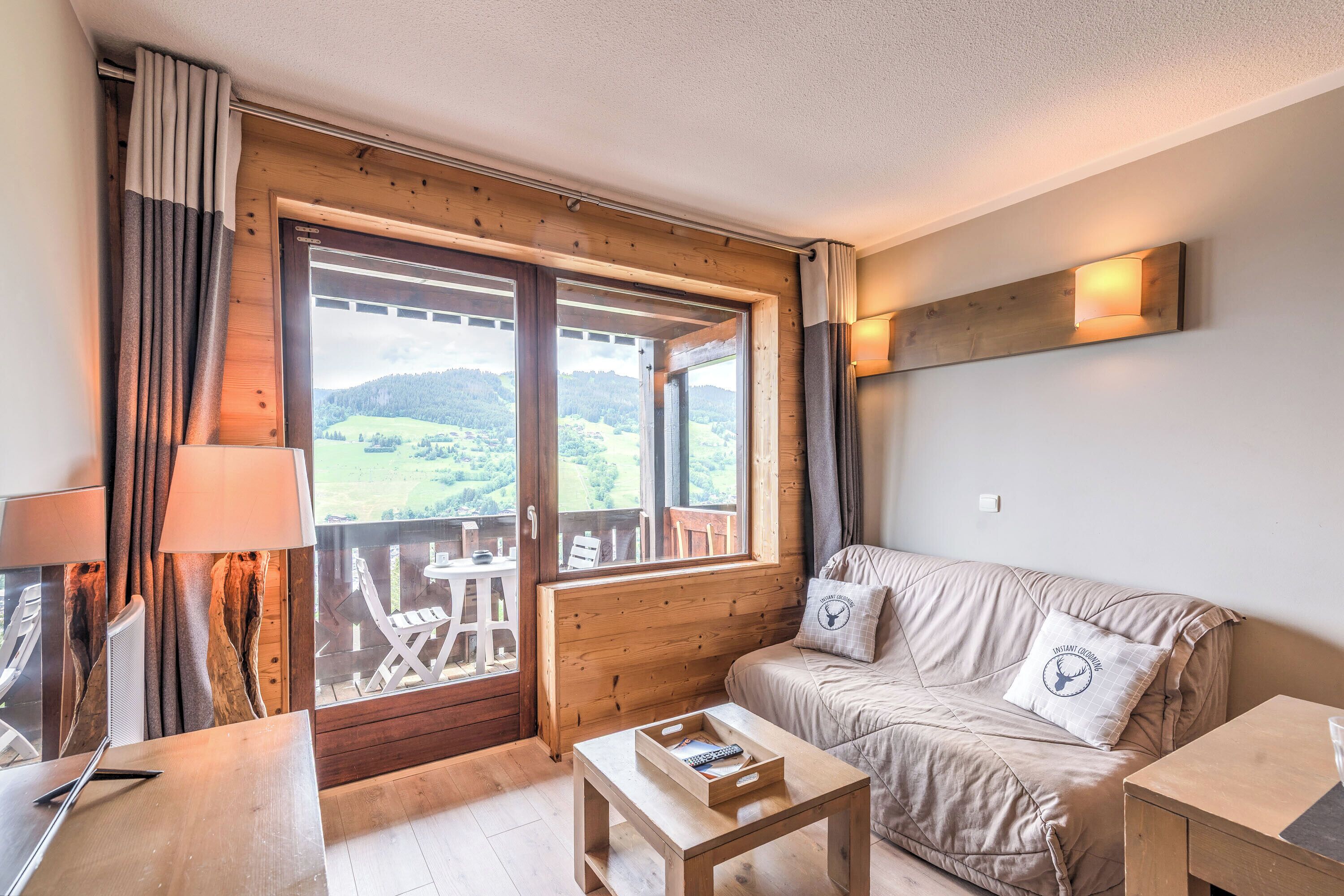 A cozy room with a wooden floor, a sofa with cushions, a small table, a lamp, and a window with a view of the outdoors.