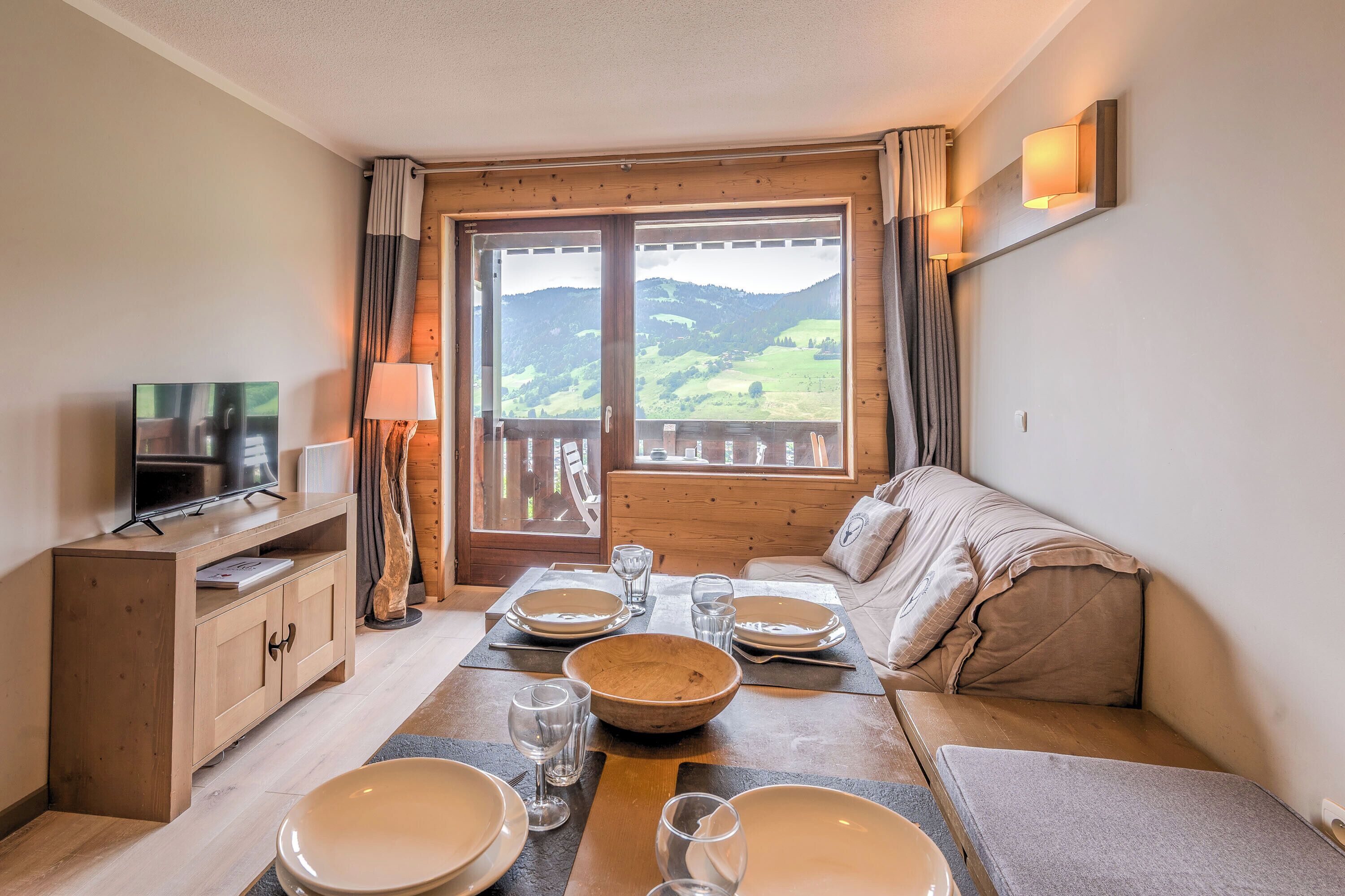 A room with a wooden table set for dining, a sofa, a TV, and a view of a mountainous landscape through a large window.