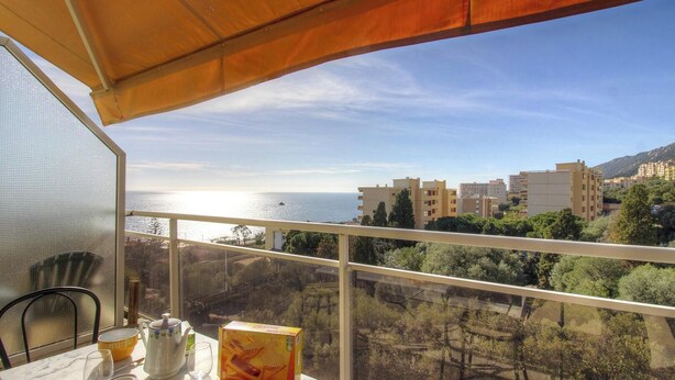 En balkon med udsigt til havet og bygninger, et bord med et tepotteskrin og en stol.