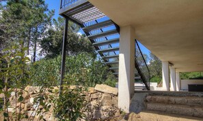 Eine Treppe mit Metallgeländern führt zu einem überdachten Bereich, umgeben von Steinmauern und Grünpflanzen.