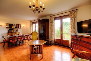 Ein Essbereich mit Holztisch und Stühlen, einer Chandelüster, einem Fernseher und einem Balkon mit Vorhängen.