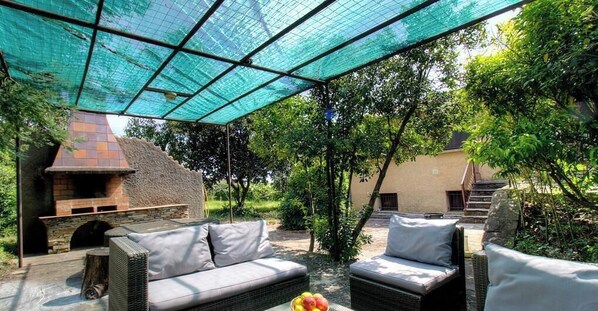 Een terras buiten met een stenen open haard, rieten meubels en een overdekt gedeelte met een blauw net.