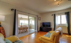 Ein Wohnzimmer mit Sofa, einem Fernseher auf einem Schrank, einem Essbereich mit Stühlen und Tisch sowie einer Glastür, die zu einem Außenbereich führt.