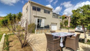 Ein zweistöckiges Haus mit Balkon, eine gepflasterte Terrasse mit Korbmöbeln und ein geparktes Auto in der Nähe.