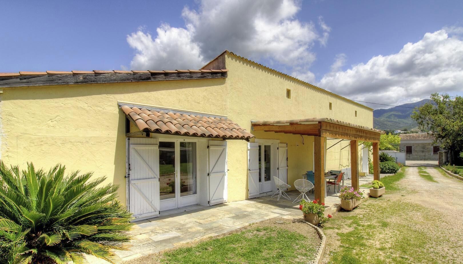 A two-story building with a tiled roof, a covered patio area with chairs, and a garden with plants.