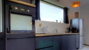 Une cuisine moderne équipée d’un four encastré, d’un four à micro-ondes et d’un réfrigérateur, avec une fenêtre agrémentée de rideaux et un luminaire suspendu.