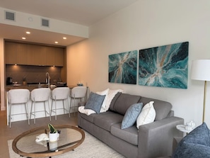 A modern living room with a grey sofa, a round coffee table, and a kitchen area with bar stools. - Lake Days & Poolside Nights (Kelowna)