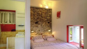 Un dormitorio con pared de piedra, cama con ropa de cama rosa, ventana con marco rojo y baño con toallero rojo.