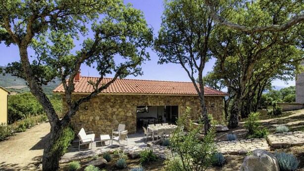 Un bâtiment en pierre avec un toit en tuiles, des places assises à l’extérieur et de la végétation environnante.