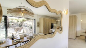 Un espace repas agrémenté d’un grand miroir, d’un luminaire suspendu et offrant une vue sur la terrasse extérieure.