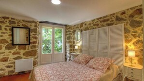 Een slaapkamer met stenen muren, een bed met bloemenbedekking, een raam met luiken en een nachtkastje met een lamp.