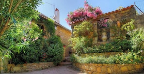 Un stretto sentiero in pietra conduce a un rustico edificio in pietra con tetto in tegole, immerso nel verde e fiorito.