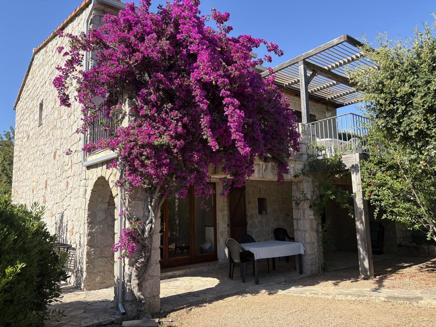 A stone building with a balcony, a table and chairs outside, and a tree with purple flowers.